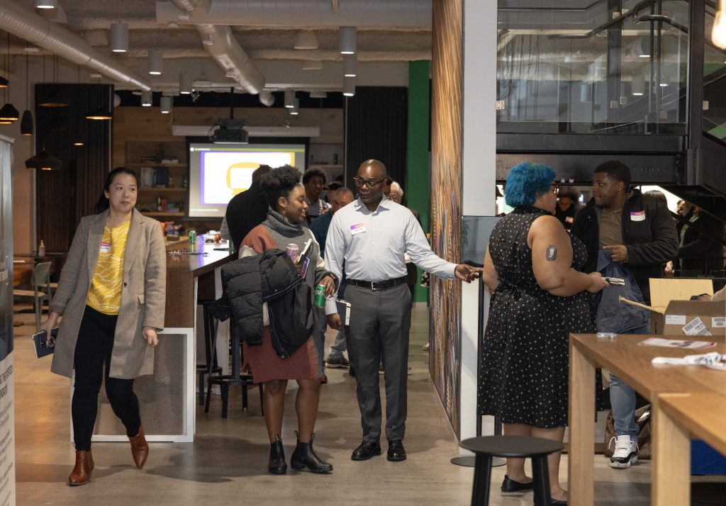 A group of people interact in a modern office space during demo day 2023; some are standing and talking while others walk through the room. Tables, a staircase, and a projector screen can be seen in this photo gallery.
