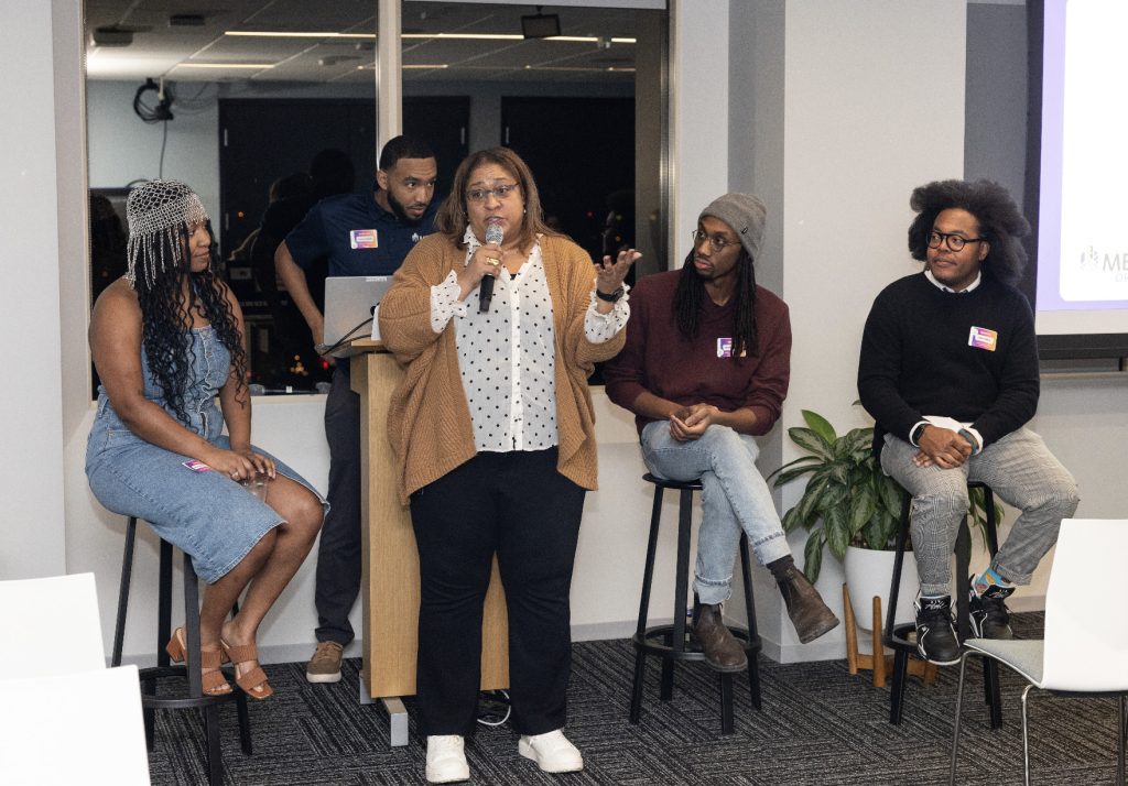 A woman speaks into a microphone while standing in front of four seated panelists in a modern room during demo day 2023. One person stands behind a podium; the panelists listen attentively. A plant and empty chairs are visible nearby.