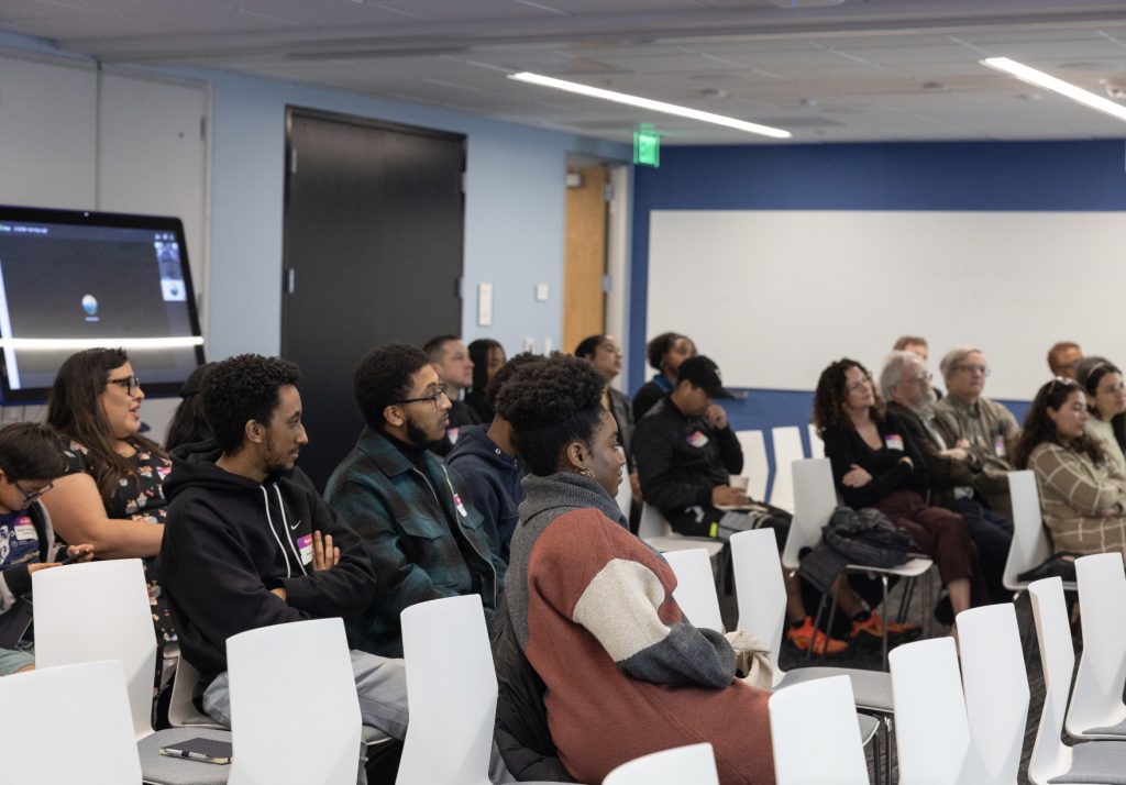 A diverse group of people sit in white chairs, attentively watching a presentation in a modern classroom with blue walls, featured in this demo day 2023 photo gallery. A screen and whiteboard are visible in the background.