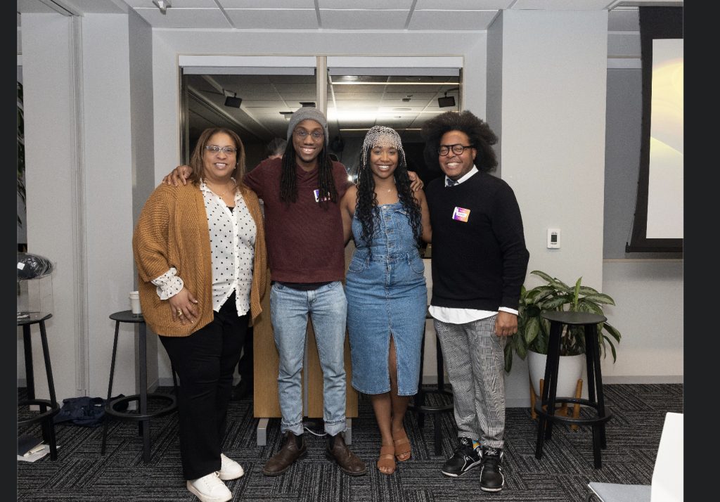 Four people stand side by side, smiling at the camera in a modern indoor setting. With plants, stools, and a presentation screen in the background, this demo day 2023 photo gallery captures a relaxed and friendly group moment.