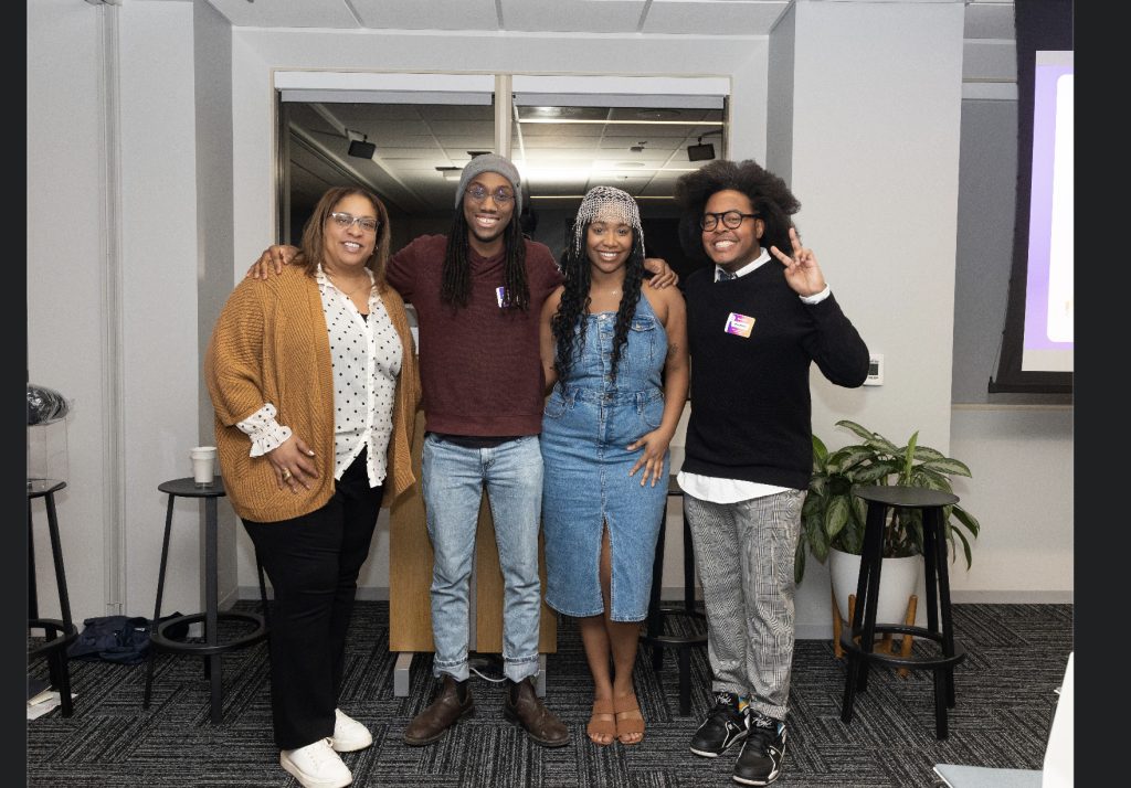 Four people stand side by side, smiling at the camera in an indoor setting for the demo day 2023 photo gallery. Three are close together; one flashes a peace sign. Stools, a plant, and a window can be seen in the background.