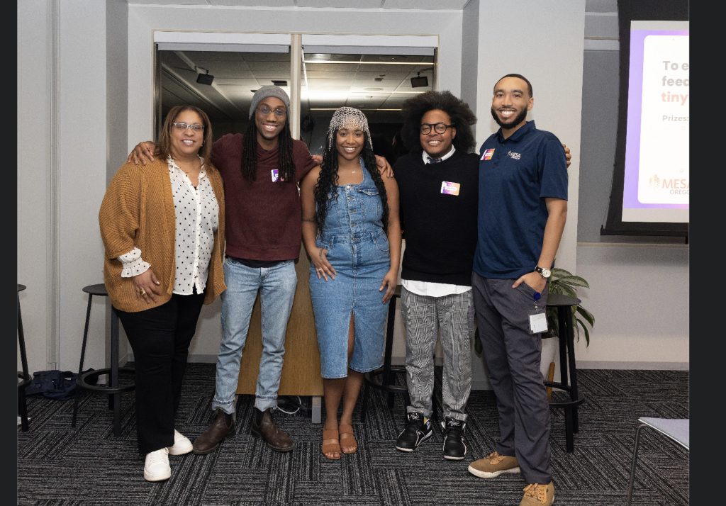 Five people stand together indoors, smiling at the camera during demo day 2023. Dressed in casual and business-casual attire, they pose in front of a presentation screen for this photo gallery moment.