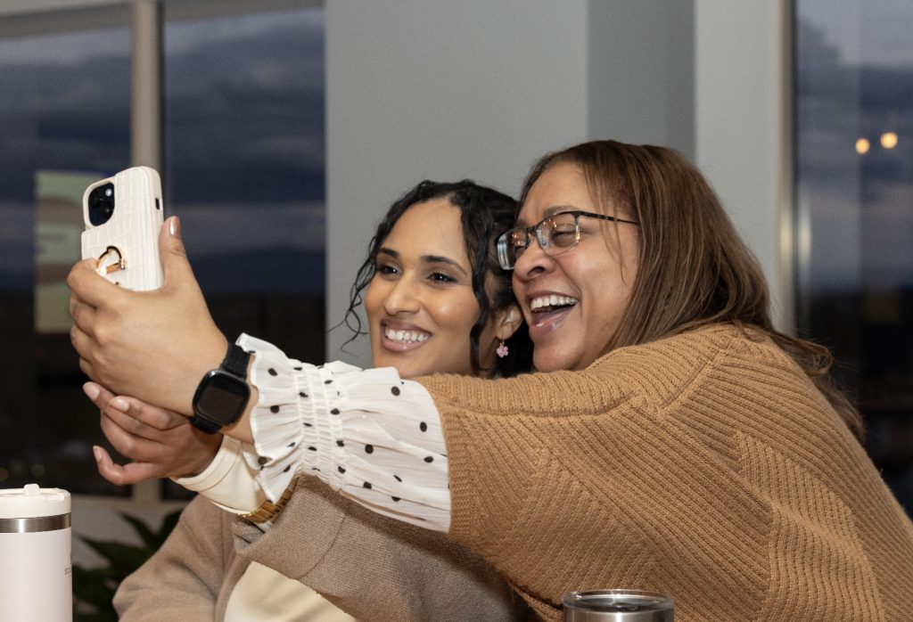 Two women smiling and taking a selfie together indoors during demo day 2023. One wears a tan sweater and glasses, the other a white blouse with black dots. Both look happy and cheerful, featured in the event's photo gallery.
