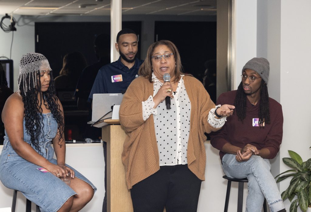 A woman speaks into a microphone while standing between three seated individuals during an indoor event; two listen attentively, and one stands behind a podium with a laptop—captured in our demo day 2023 photo gallery.