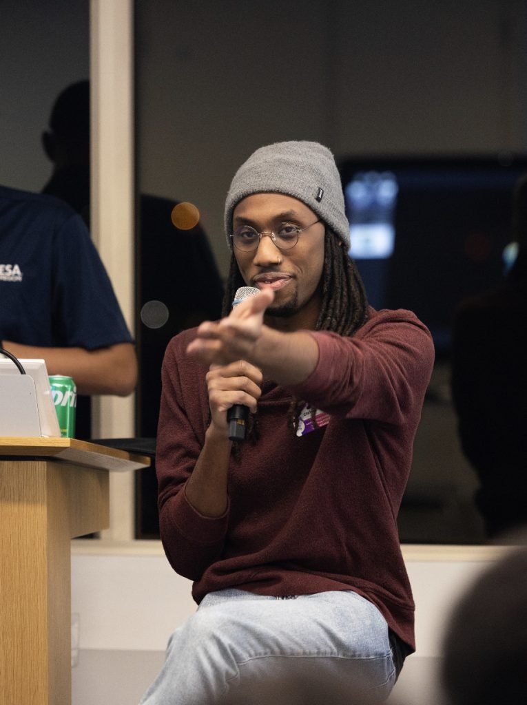 A person with long hair and a gray beanie sits on a chair, holding a microphone and gesturing forward with their hand. They wear glasses, a maroon shirt, and jeans. A laptop and drink can are on a podium nearby in this demo day 2023 photo gallery.