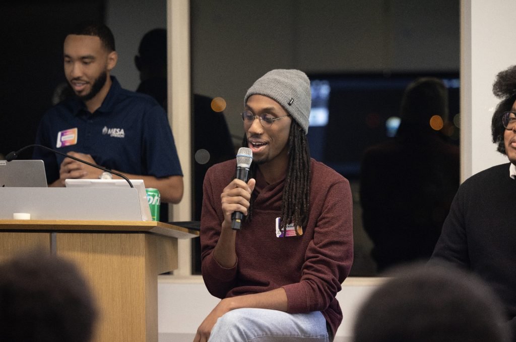 A person wearing a gray beanie speaks into a microphone while seated, as another stands at a podium with a laptop nearby. The setting appears to be an indoor event, captured in the demo day 2023 photo gallery.