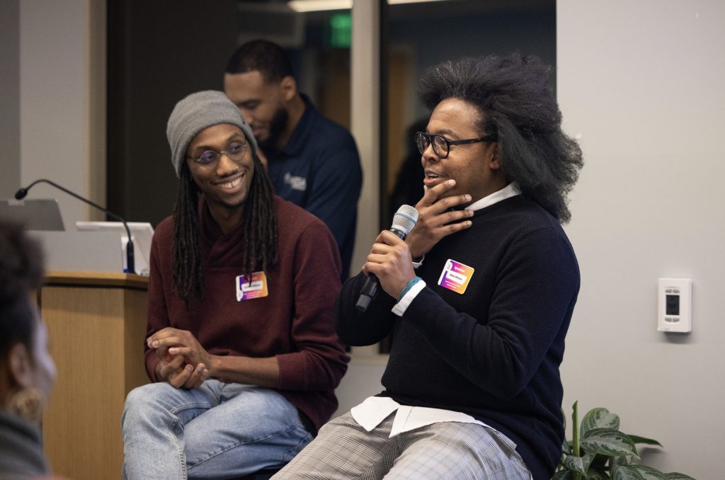 Two people sit and talk during a panel discussion at Demo Day 2023, one holding a microphone and speaking while the other smiles. Both wear conference name tags. A third person is in the background by a podium. See more in our photo gallery.