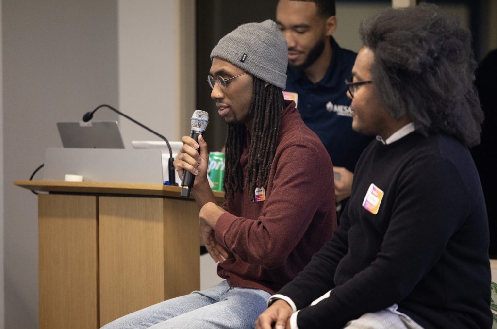 Two men sit and speak at an indoor event—one holding a microphone while the other listens. Both wear nametags, with a podium, laptop, and microphone in the background, captured in this Demo Day 2023 photo gallery.