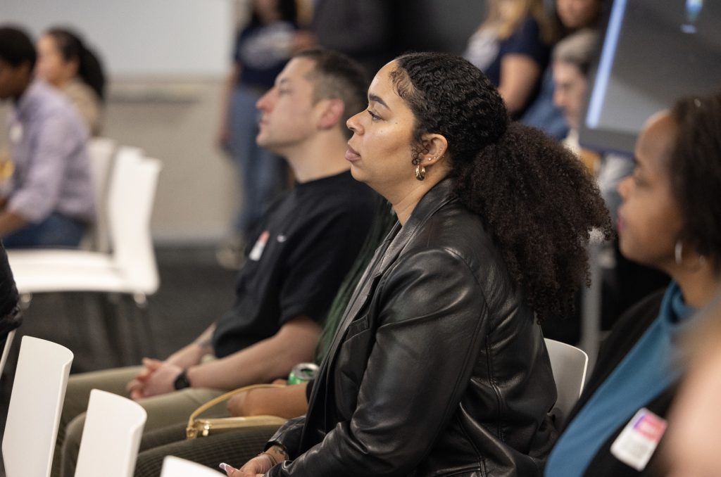 A woman with curly hair in a ponytail and a black leather jacket sits attentively in a conference room among other seated people, all facing forward and listening in this demo day 2023 photo gallery.