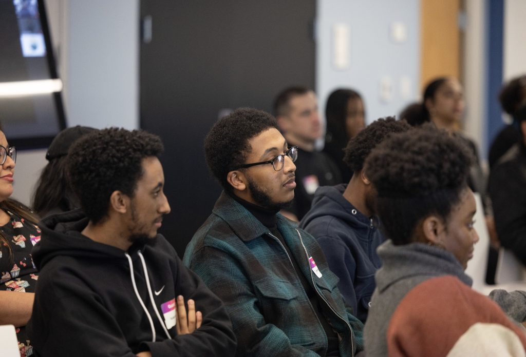 Several people sit closely together in a classroom or seminar setting during demo day 2023, attentively listening to a speaker. Most are facing forward, wearing casual clothes, and some have name tags on their shirts for the photo gallery.
