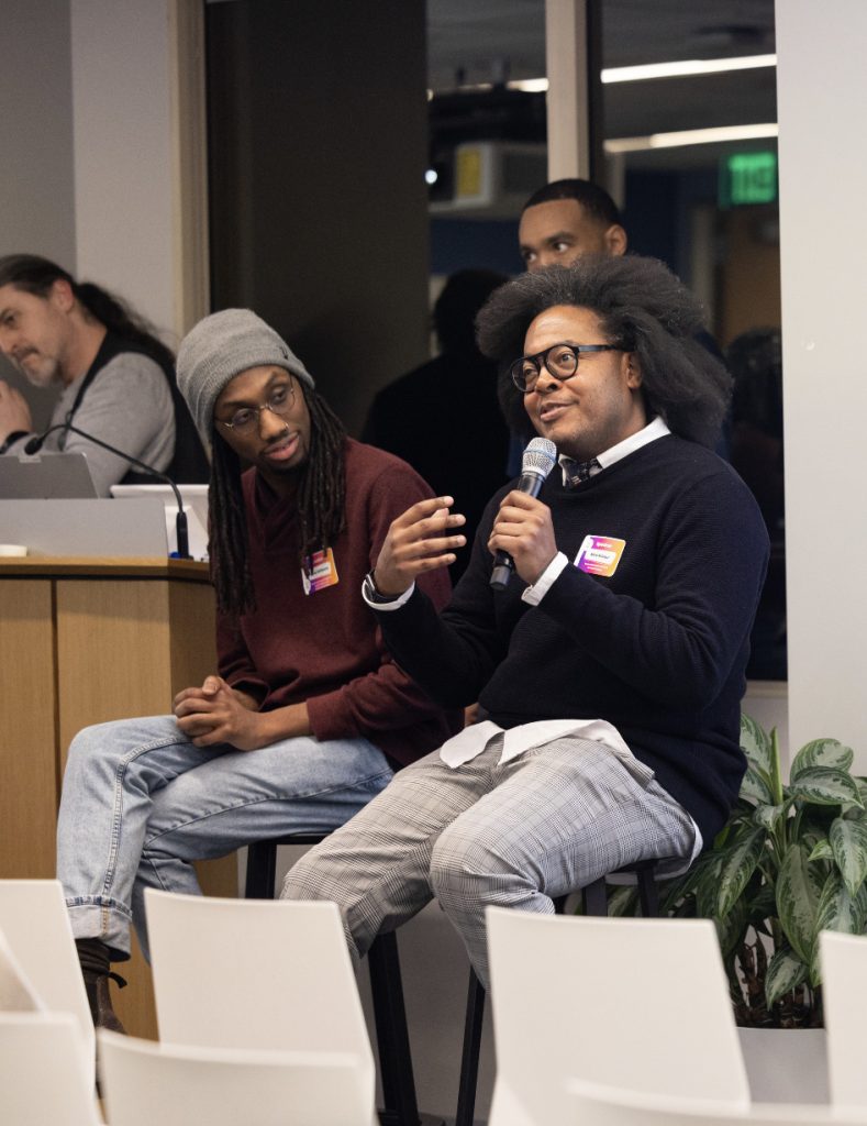 Two men sit at the front of a room during the Demo Day 2023 photo gallery; one speaks into a microphone while gesturing, and the other listens attentively. Both wear glasses and conference name tags, with several people and a plant in the background.