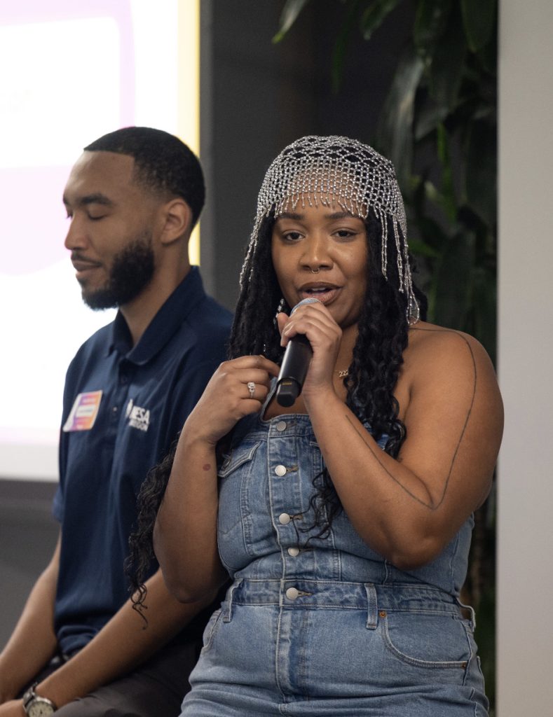 A woman in a jeweled headpiece and denim outfit speaks into a microphone while seated at demo day 2023. A man in a navy shirt sits beside her with his eyes closed. A plant and the blurred screen complete this photo gallery moment.