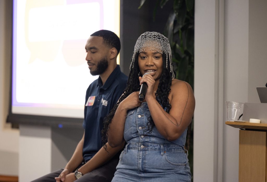 A woman in a denim outfit speaks into a microphone while sitting next to a man with his eyes closed. Behind them, a presentation screen hints at the event—part of the demo day 2023 photo gallery.