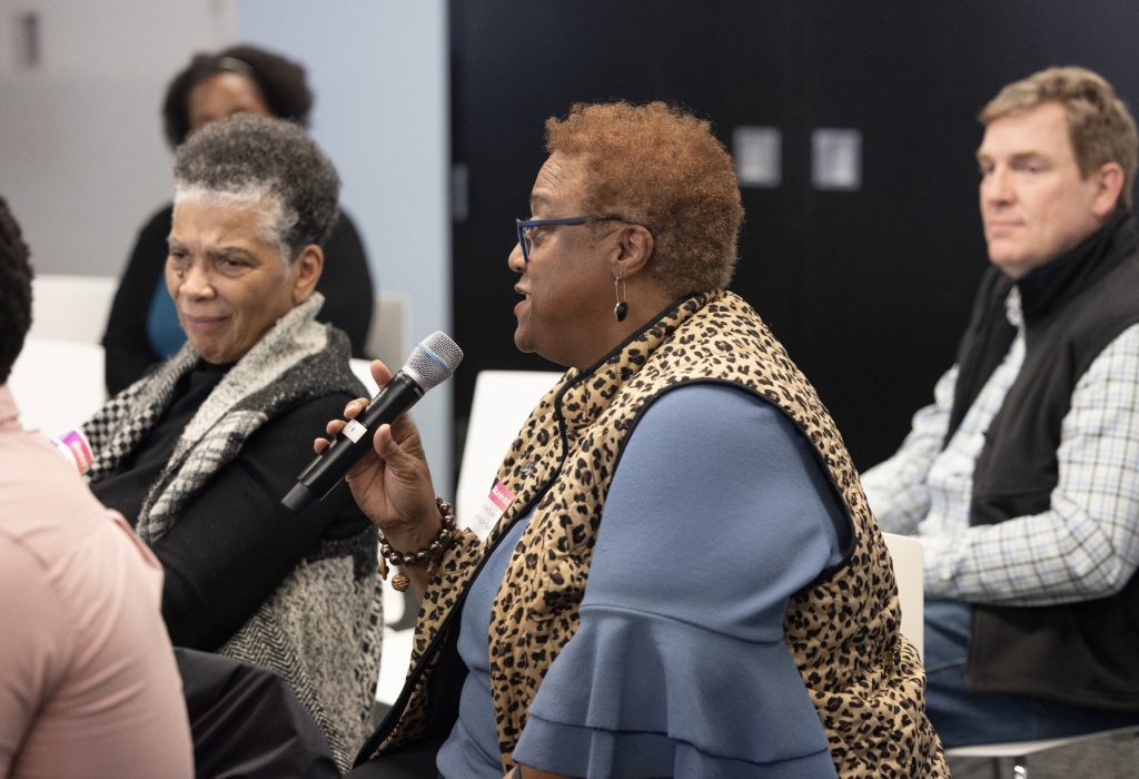 A woman wearing glasses and a leopard-print vest speaks into a microphone during a meeting at demo day 2023, while others sit and listen attentively in the background. Explore more moments like this in our photo gallery.