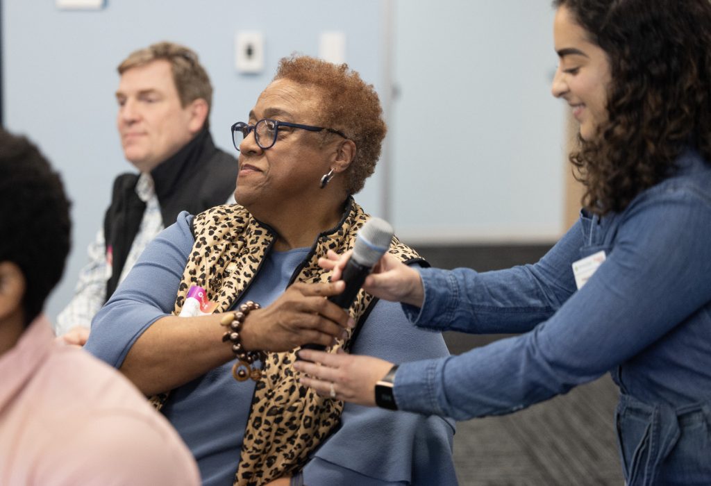 A woman wearing glasses and a leopard-print scarf receives a microphone from a smiling woman in a denim jacket during an indoor demo day 2023 event. Other attendees are visible in the background of this photo gallery.