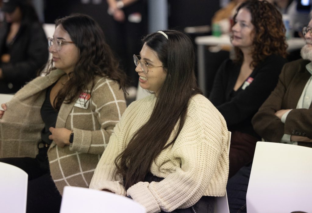 Several people sit indoors, attentively listening to something off camera. Two women in the foreground wear glasses and cozy sweaters, while others are visible in the background in this casual, relaxed demo day 2023 photo gallery.