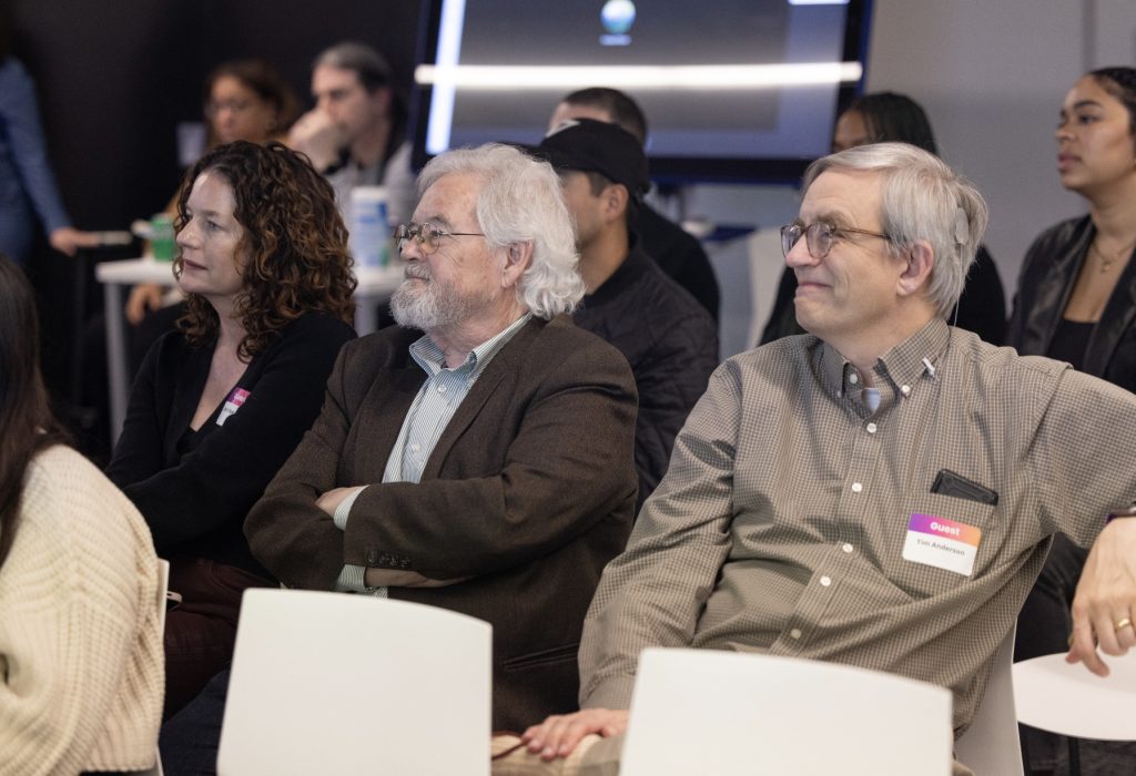 A group of people sit in white chairs, attentively watching something off-camera during demo day 2023. Three individuals in the front row appear focused, while others are seated and standing in the background. Explore more in our photo gallery.