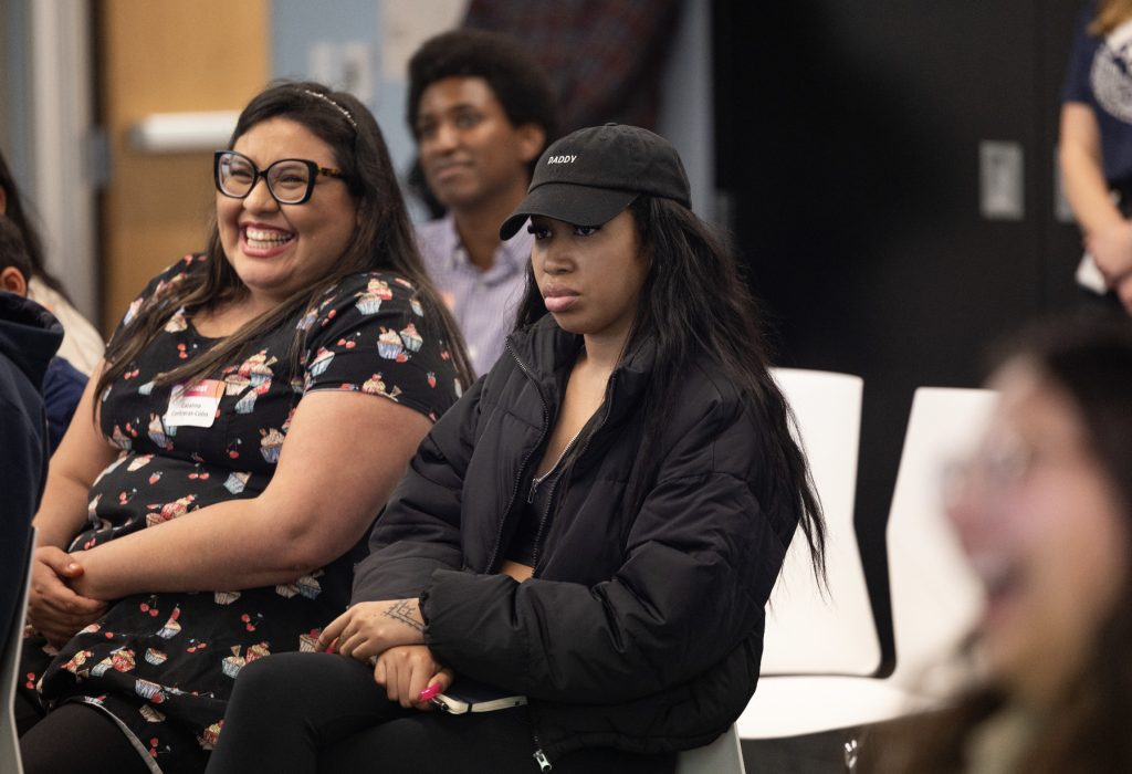 Two women sit side by side at an indoor event in this demo day 2023 photo gallery; one is smiling and laughing, while the other wears a black cap and jacket, looking serious. Other attendees are visible in the background.