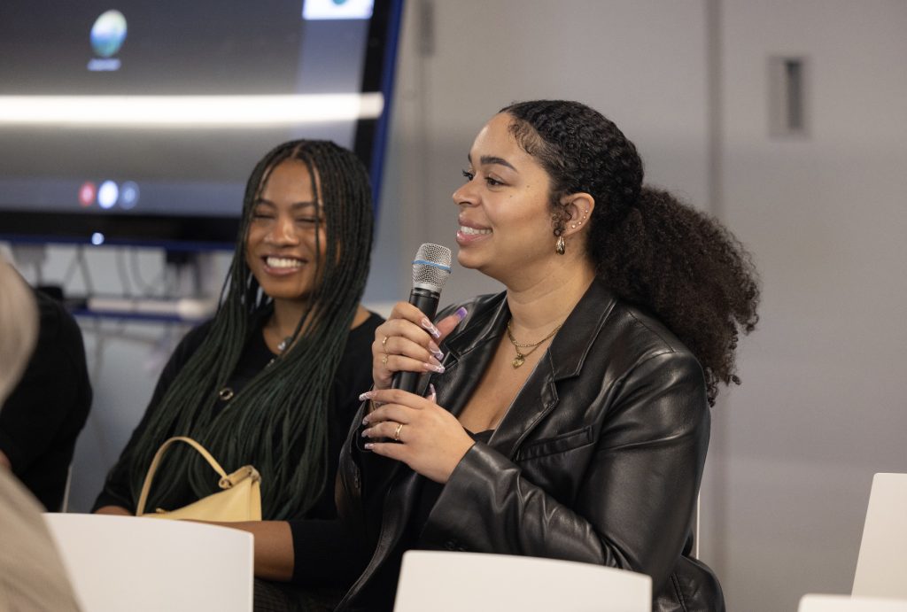 Two women sitting and smiling indoors; one in a black leather jacket speaks into a microphone as the other, with long braided hair, listens. A large screen glows in the background at this lively demo day 2023 photo gallery moment.