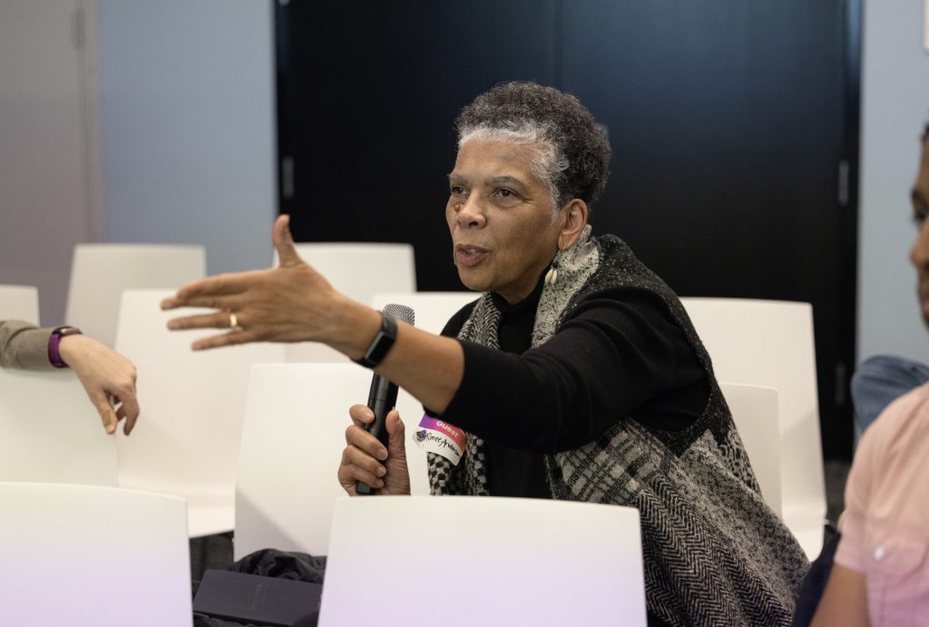 An older woman sits in a chair, holding a microphone and gesturing with her left hand while speaking during demo day 2023. She is wearing a black top and patterned scarf, with empty white chairs behind her in this photo gallery.