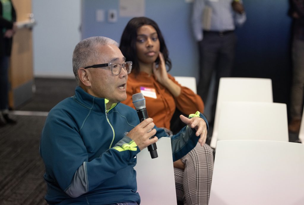 A man wearing glasses and a blue jacket speaks into a microphone while seated in a row of white chairs at the demo day 2023 photo gallery. A woman in an orange top and plaid pants sits behind him, listening while others stand in the background.