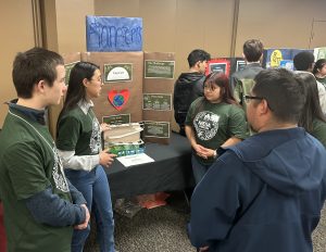 A group of students at a science fair stand near a display board labeled “Pioneers.” It’s a Wrap on our 2023 Demo Days! One student explains the project, while the table holds model materials and posters. Everyone is engaged and listening.