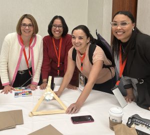 Four women smiling around a table, participating in a group activity indoors. With conference lanyards on, they embody Turning Passion Into Action as they engage with a wooden triangular structure, game cards, a pink phone, and coffee cup.