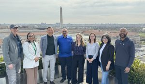 A group of eight people stand smiling on a rooftop with the Washington Monument visible in the background on a cloudy day, dressed in business casual attire—turning passion into action as they gather for an inspiring occasion.