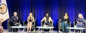 Five people are seated on stage in armchairs with microphones, participating in a Turning Passion Into Action panel discussion. They sit before dark blue curtains, with round glass tables, bottled water, and a flag to the left.