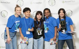 Five smiling students wearing medals and blue shirts pose together, holding awards, books, and a plaque in front of an Oregon State University and Oregon MESA backdrop—captured for the Mesa Day 2024 photo gallery.