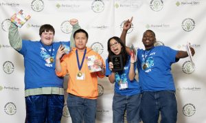 Four smiling students stand in front of a Portland State University backdrop at Mesa Day 2024, wearing medals and holding prizes. Three wear blue shirts, one an orange hoodie; one holds a plaque. They gesture excitedly toward the camera for the photo gallery.