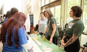 A group of people interact at an indoor informational booth with brochures, buttons, and pencils on a green table. Two women in green shirts stand behind the table smiling, while others engage and explore the Mesa Day 2024 photo gallery materials.