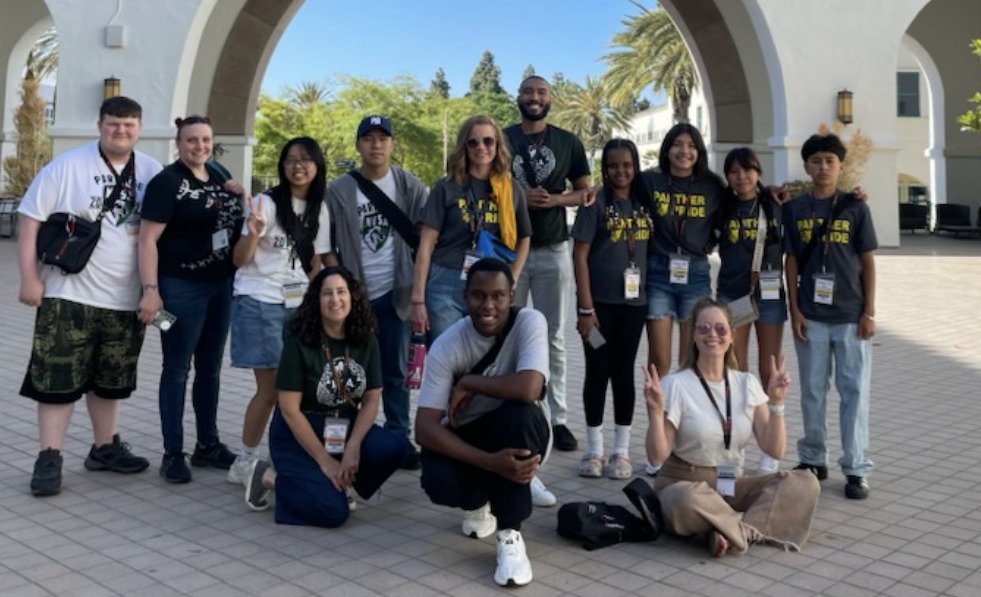 A group of thirteen people, mostly young adults and teens, pose and smile together outdoors under large arches after the National Engineering and Design Competition. Palm trees and buildings are visible in the background.