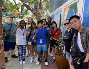 A diverse group of teens and two adults pose together, some making peace signs, outside in front of colorful murals and festive banners at the National Engineering and Design Competition, enjoying a sunny day.