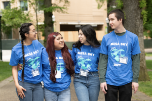 Four students wearing matching blue MESA DAY 2024 shirts and name badges stand outdoors, smiling and chatting with arms around each other, celebrating the EXPANSION of their program in a green, tree-filled campus setting.
