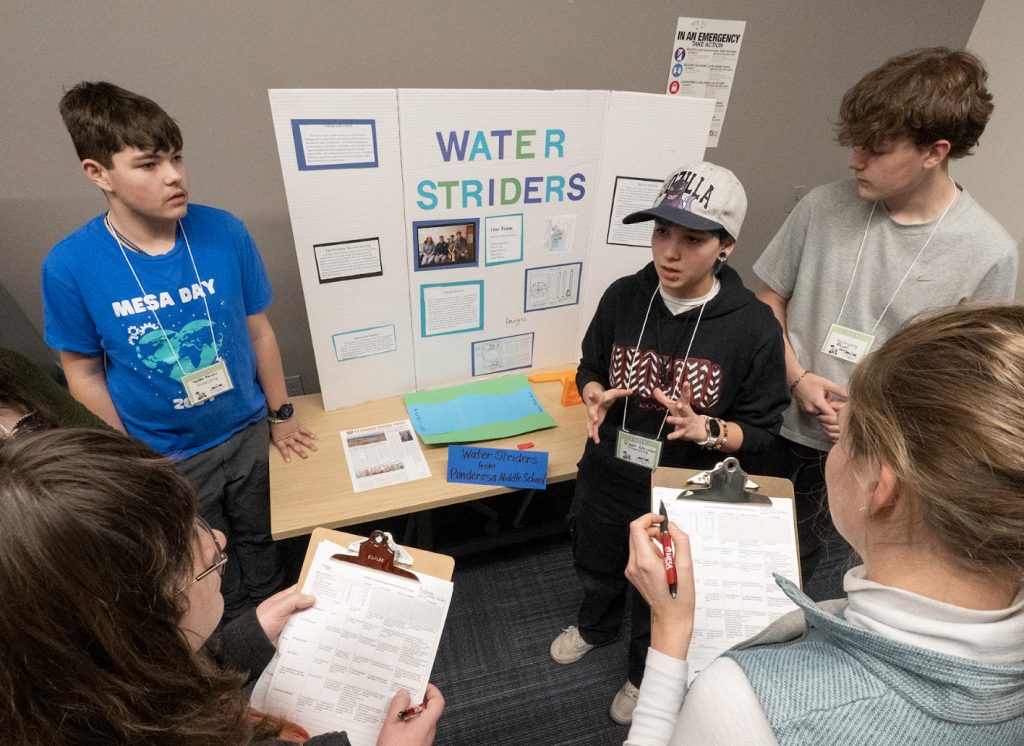 A group of students stands around a display board titled Water Striders, discussing their project with judges holding clipboards in a classroom setting for the Demo Day 2025 photo gallery.