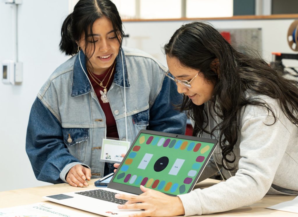 Two young women smile and look at a laptop with a colorful board game displayed on the screen. One stands in a denim jacket; the other, seated and wearing glasses, reaches toward the laptop. Perfect for the demo day 2025 photo gallery.