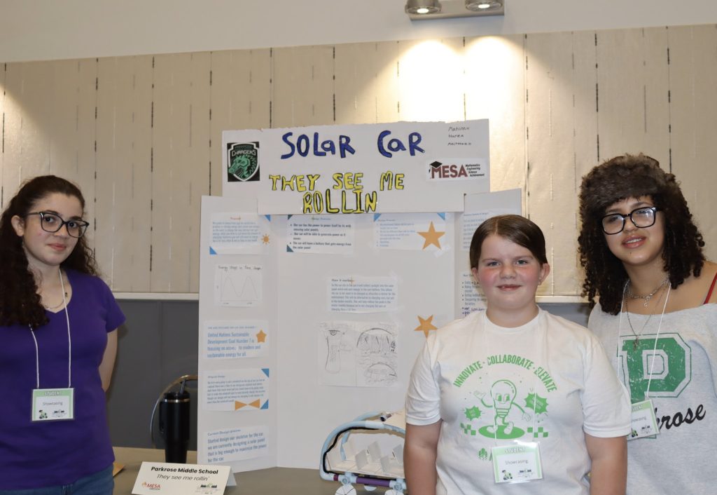Three students smile in front of their science fair project, “Solar Car: They See Me Rollin,” featuring diagrams and star decorations. This moment is part of the demo day 2025 photo gallery, with name tags visible on each student.