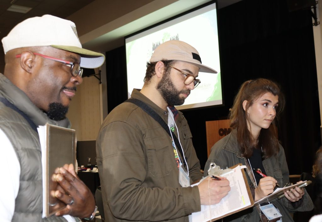 Three people stand indoors holding clipboards and writing at demo day 2025. Two wear beige caps and glasses, one in a white vest, the third in a dark jacket. A projector screen is visible in the background for the photo gallery.