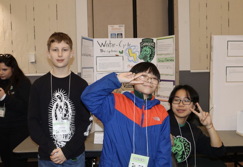 Three students stand in front of a science fair project display about the water cycle at demo day 2025. Two flash peace signs and smile, while the third stands with hands by their sides. Other displays and people appear in the background for the photo gallery.
