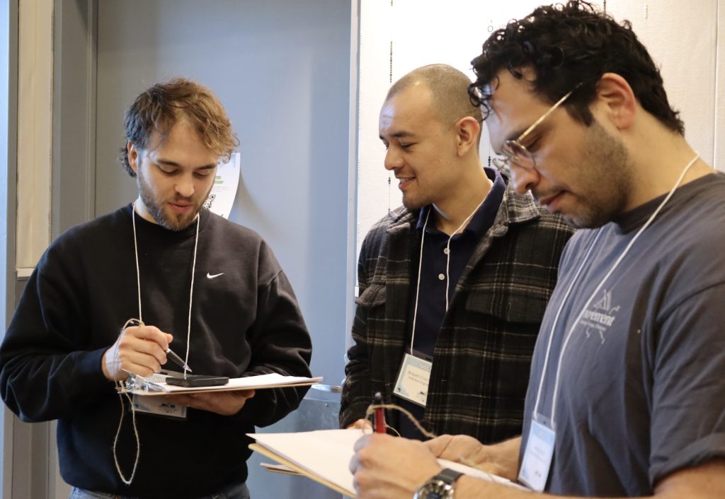 Three men wearing name badges and casual clothes stand together indoors. Two are holding clipboards and pens, writing or taking notes, while the third man smiles and looks on—a candid moment from our demo day 2025 photo gallery.