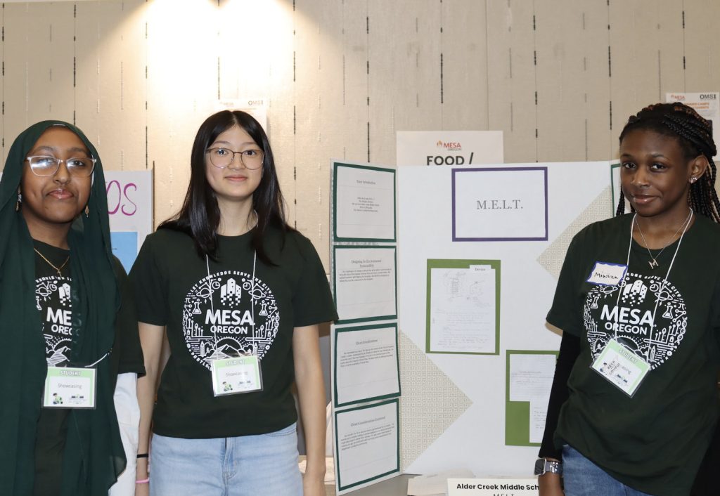 Three students stand in front of a science fair display board labeled “M.E.L.T.” for the Demo Day 2025 photo gallery. They wear matching green MESA Oregon T-shirts and name tags, with project papers displayed on the board.