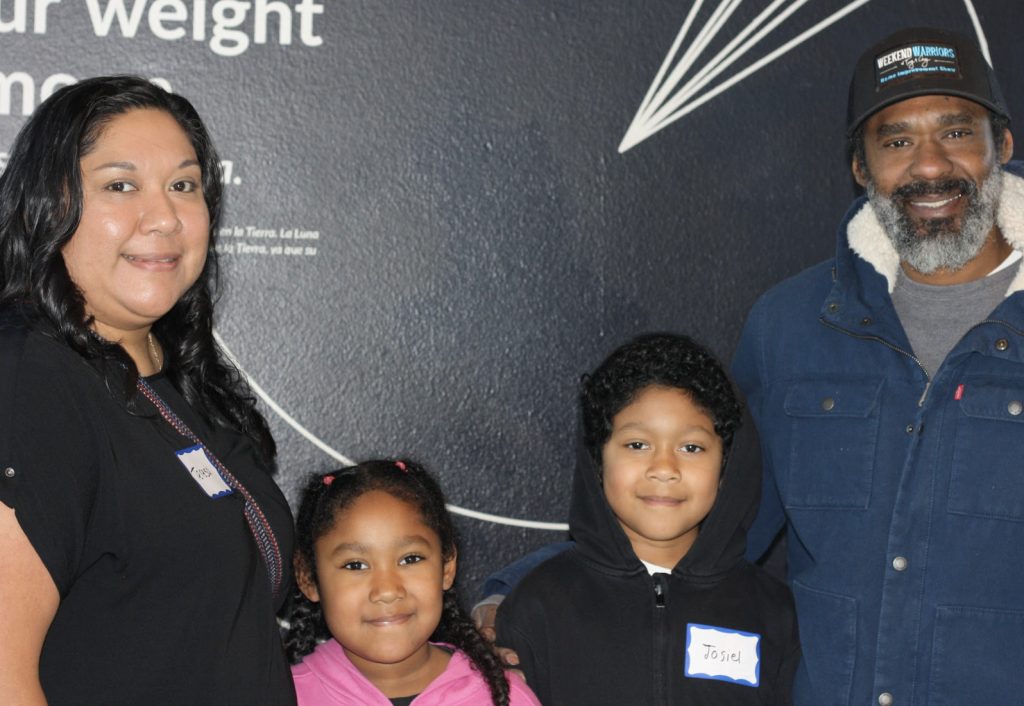 A smiling group of four people, two adults and two children, stand together in front of a dark wall for the demo day 2025 photo gallery. The adults are on the ends, and the children are in the middle, all wearing name tags.