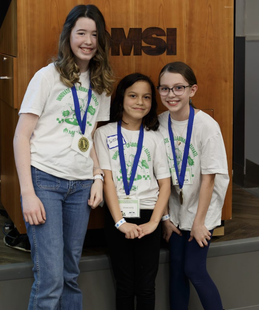 Three girls wearing matching white t-shirts with green text and blue ribbon medals pose and smile in front of a wooden podium with MSI written on it. This demo day 2025 photo gallery snapshot shows two girls with glasses, all sporting name tags.