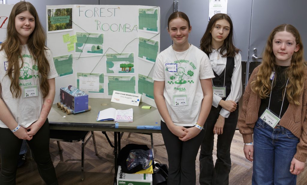 Four girls stand in front of a science fair display titled Forest Roomba, featuring posters, drawings, and a small model on a table. Two wear event t-shirts, all have name tags—part of the demo day 2025 photo gallery.