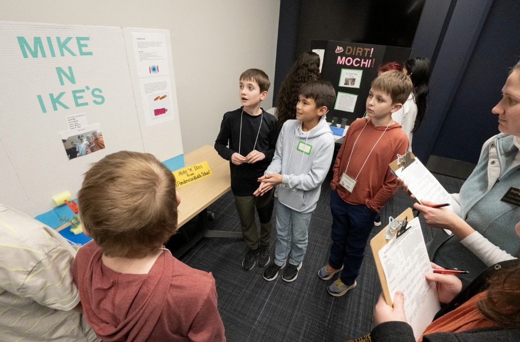 A group of children stand in front of a presentation board labeled MIKE N IKE’S while others observe and take notes on clipboards at demo day 2025, creating moments perfect for the event’s photo gallery.