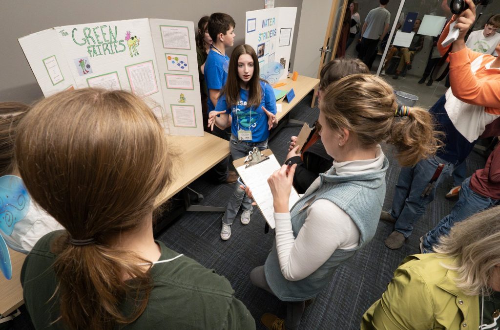Three students present their science project called Green Fairies to two judges with clipboards during demo day 2025, while other groups and posters are visible in the background of a busy classroom for the photo gallery.
