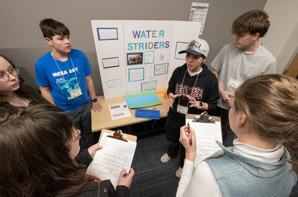 Six students stand around a table with a display board titled Water Striders. One gestures while others listen and hold clipboards, appearing to present their project for the Demo Day 2025 photo gallery in a classroom setting.