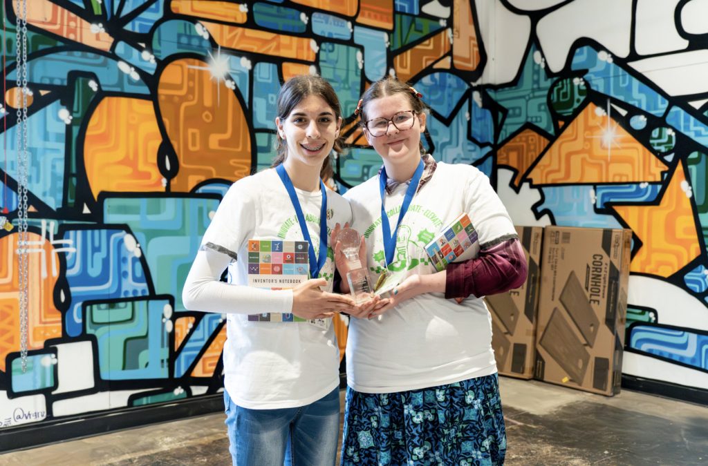 Two people wearing white T-shirts and blue lanyards stand smiling together, holding a trophy in front of a colorful, abstract mural. Cardboard boxes are visible in the background—part of our demo day 2025 photo gallery.