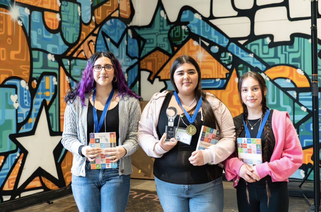 Three girls stand side by side in front of a colorful, geometric mural, wearing medals and holding books and trophies, smiling at the camera for the Demo Day 2025 photo gallery.